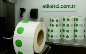Rulo etiket üreticisi olan Etiketci.com.tr üretim tesisinde, barkod cihazlarının yanında duran ve üzerinde yeşil yuvarlak etiketler bulunan büyük rulo etiketler.